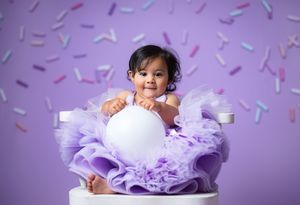 A baby in a lavender tutu dress lies on a white floor with confetti sprinkled around in a dreamy portrait setting.