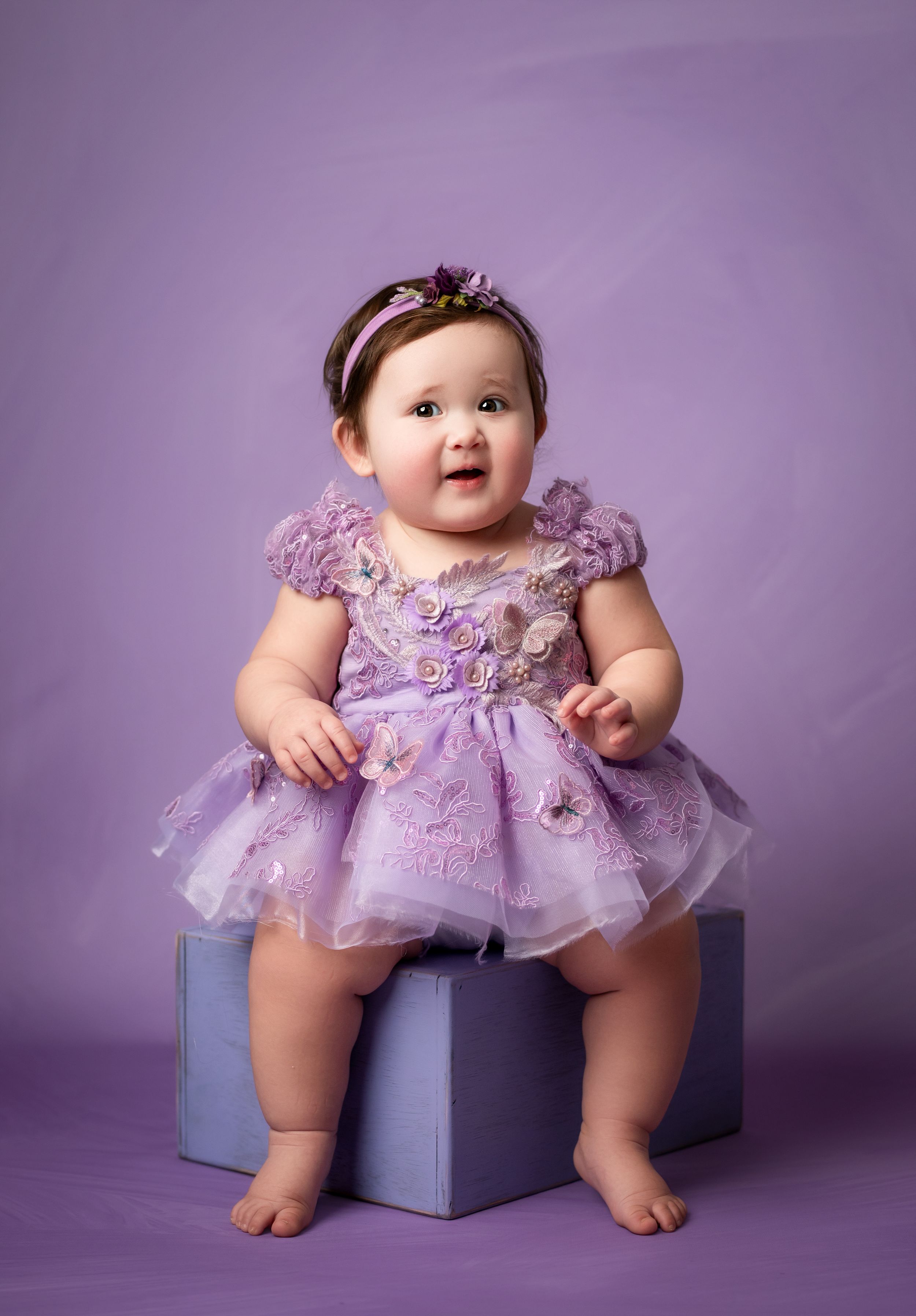 Baby in purple tulle dress sits on blue cube against purple backdrop in professional studio portrait series.