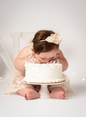 A baby wearing a peach floral headband lies face down in front of a white frosted cake in a studio setting.