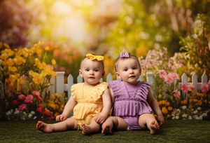 Two baby girls in yellow and purple outfits sit together in a colorful flower garden with a white picket fence backdrop.