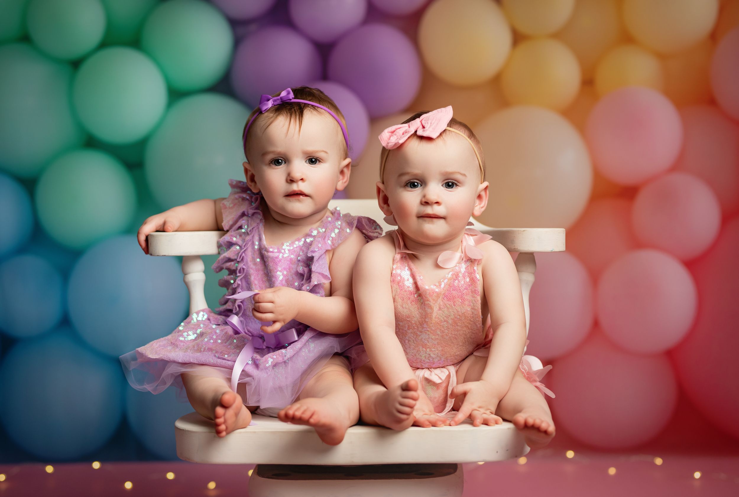 Two baby girls in pink and purple dresses sit together in front of a colorful rainbow balloon backdrop.