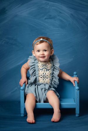 Smiling baby girl in blue ruffled romper sitting on a small blue chair against a blue backdrop.