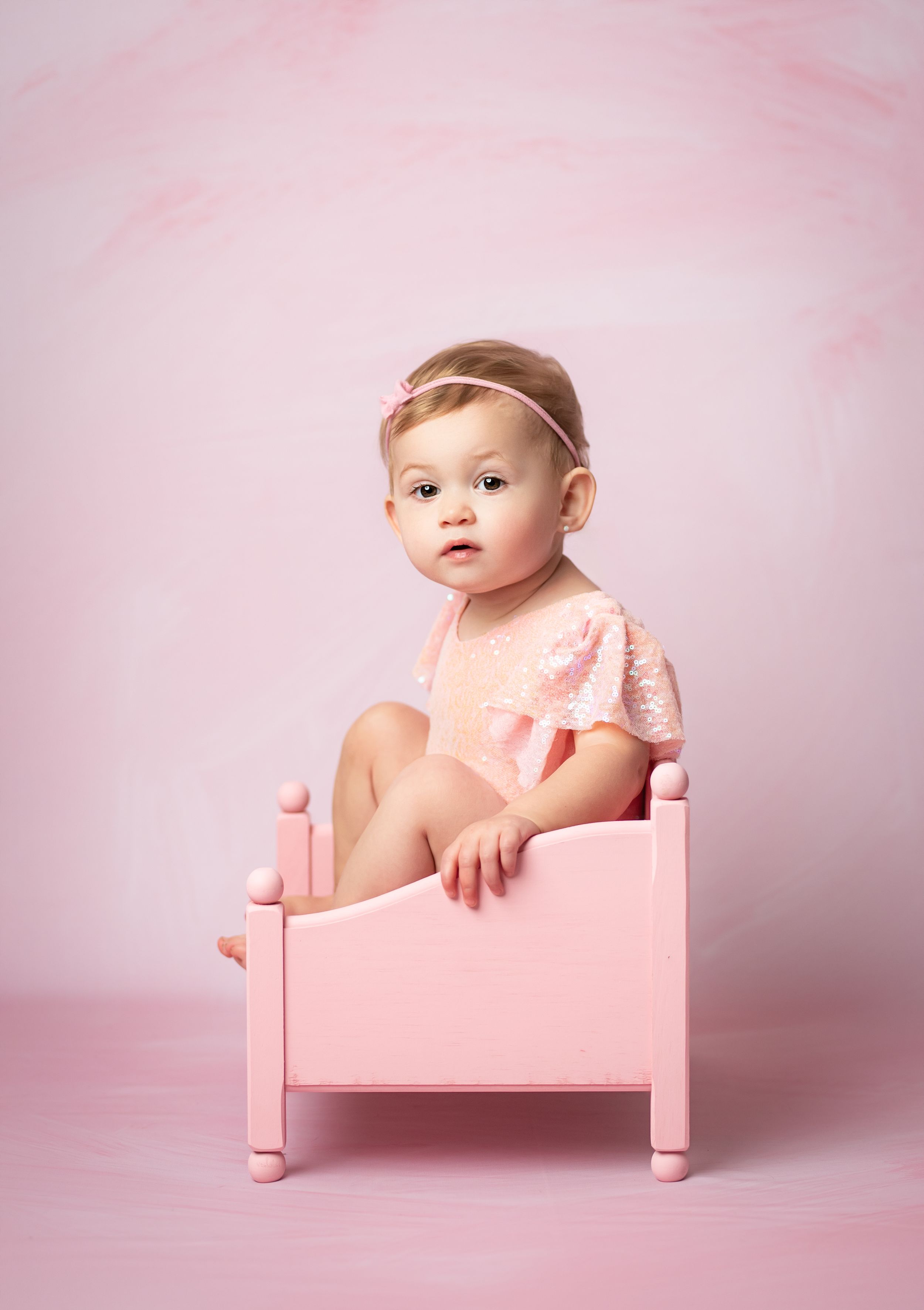A baby girl in a pink lace outfit sits in a small pink wooden bed against a soft pink background.