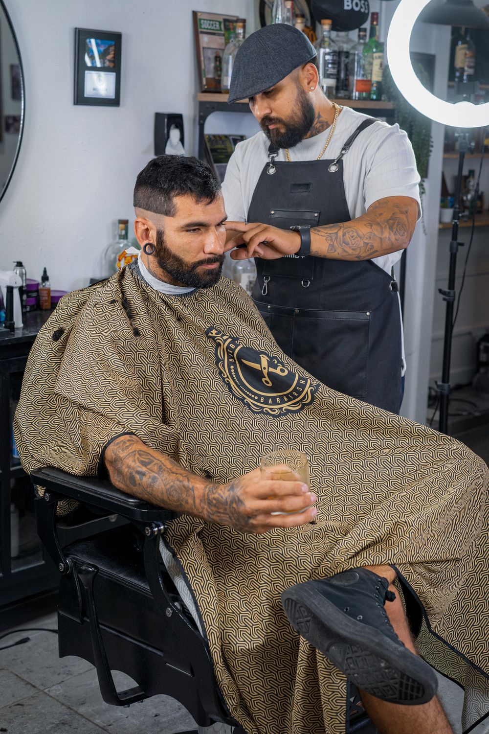 Bearded client holding drink while barber styles hair in modern barbershop.
