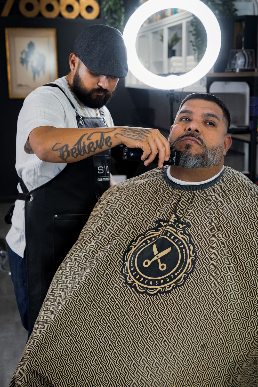 Tattooed barber grooming bearded client under ring light in dark barbershop.