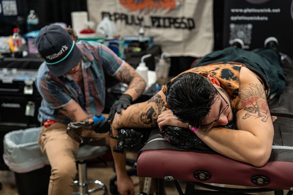 Tattoo artist working on back tattoo of seated client at convention booth.