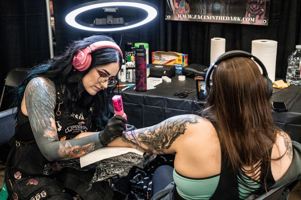 Female tattoo artist wearing headphones inks a detailed tattoo on a client's arm at a tattoo convention.