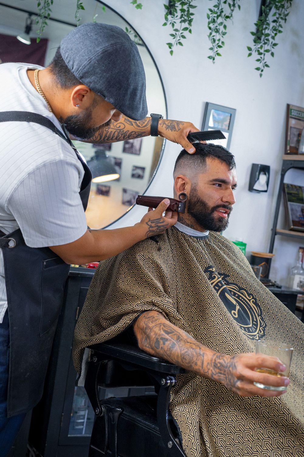 Tattooed barber using clippers on client holding drink in stylish barbershop.