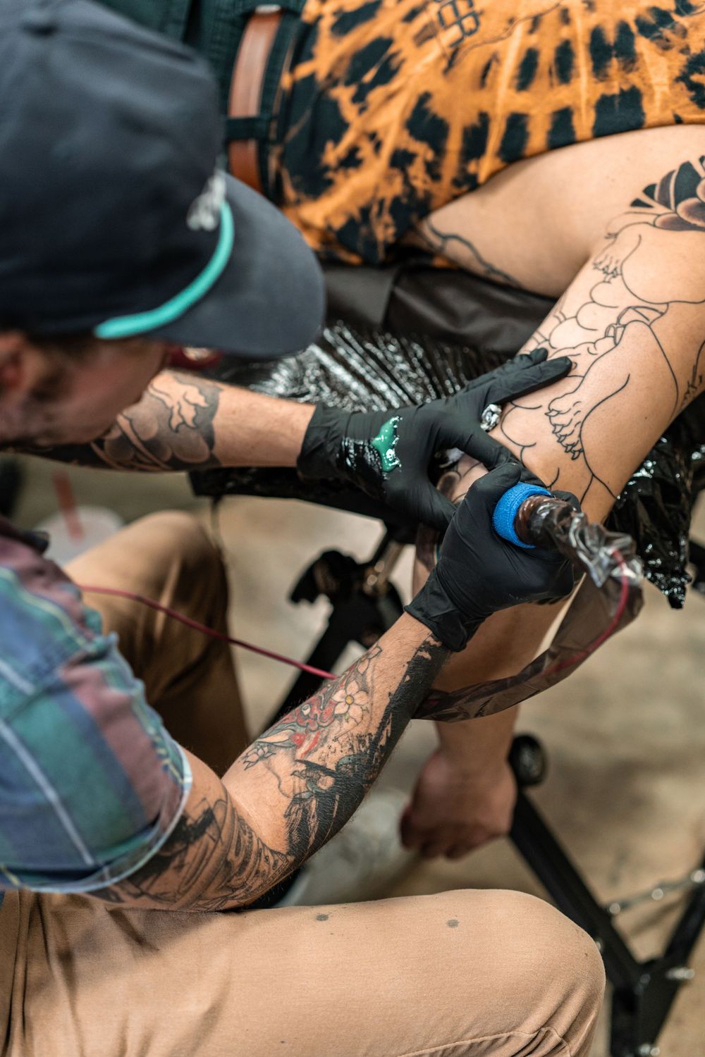 Heavily tattooed artist wearing black gloves carefully applies a tattoo on a client's leg using a tattoo machine.