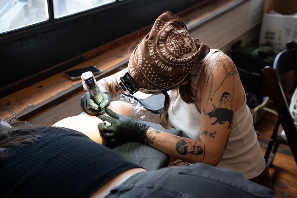 Tattoo artist wearing a decorated hat and mask tattooing a client in a professional studio.