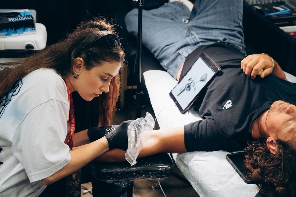 Female tattoo artist carefully inking client's arm at outdoor tattoo event.