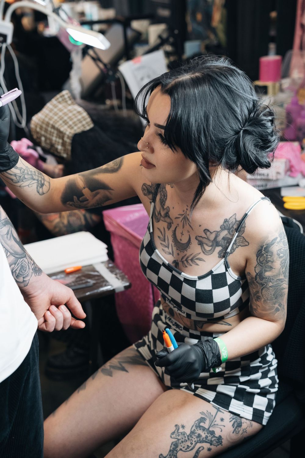 Heavily tattooed woman in checkered outfit works at a tattoo convention booth.