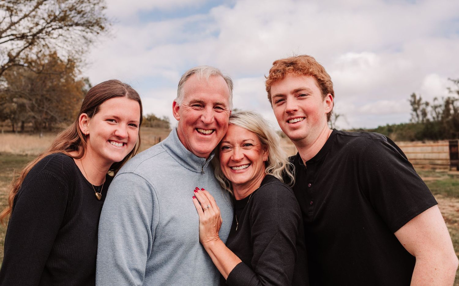 Smiling family of four posing together outdoors in open field under cloudy sky.