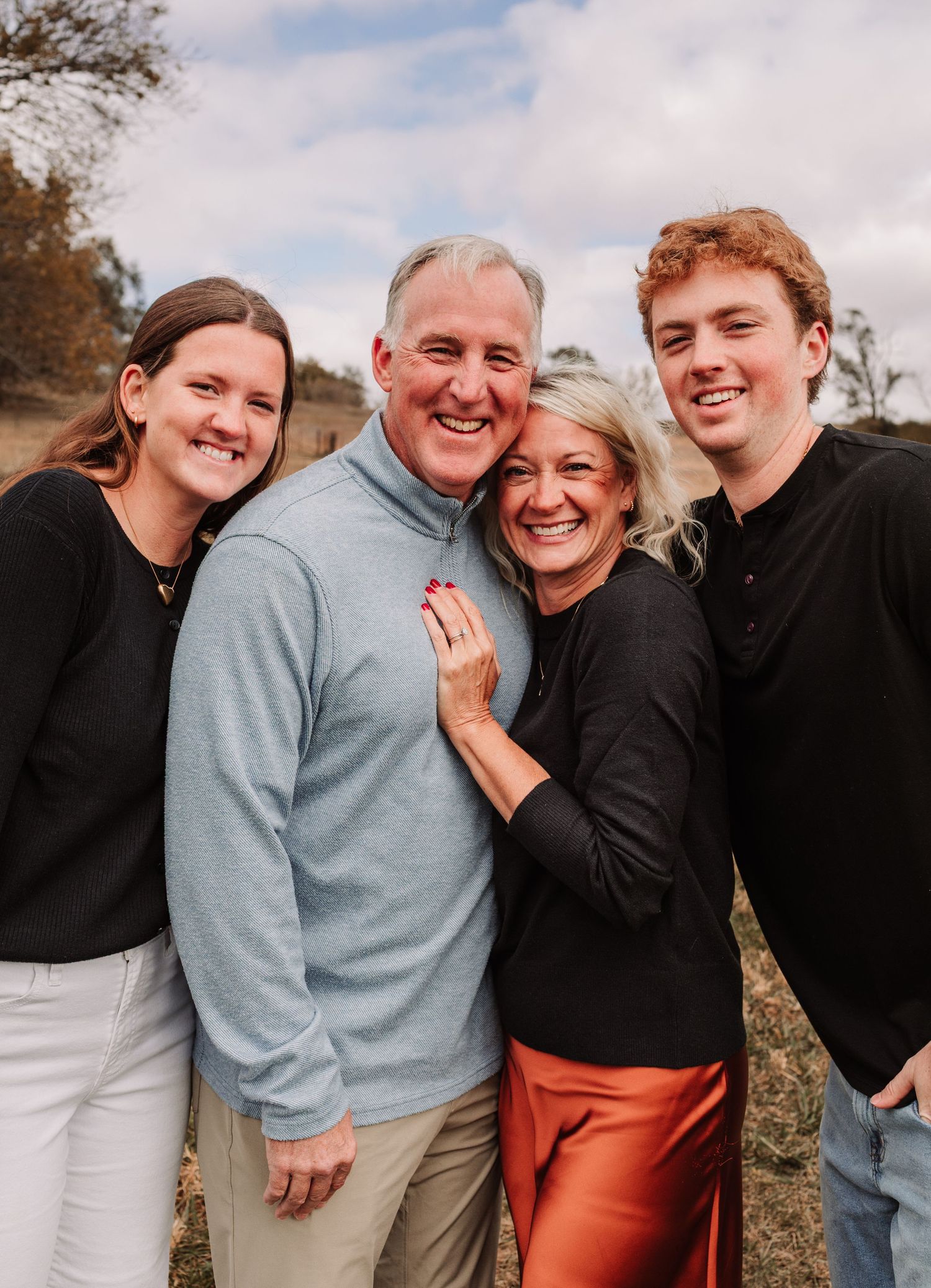 Happy family of four posing outdoors in fall setting, parents and two young adult children smiling warmly.