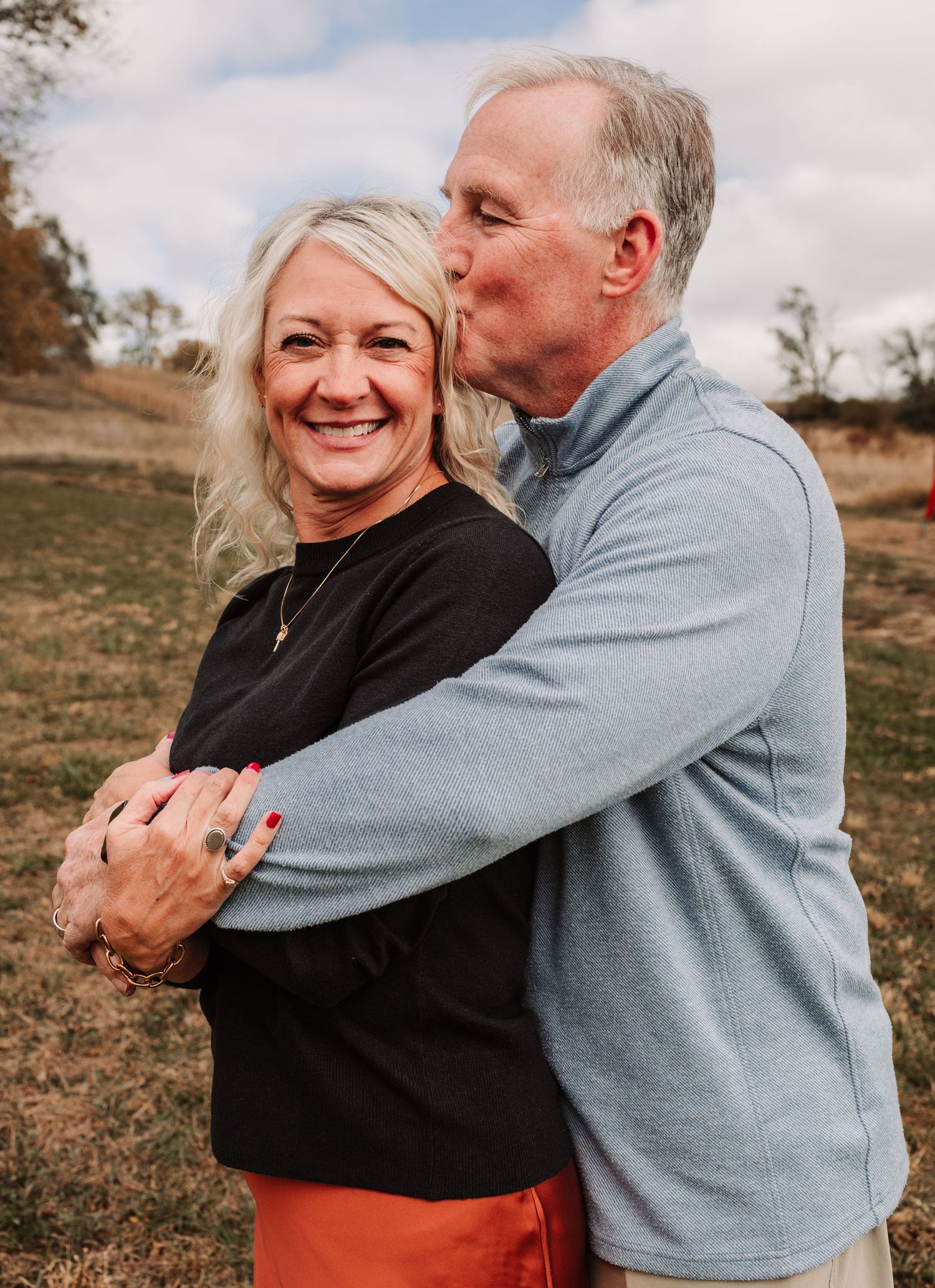 Loving couple embracing outdoors, man in blue fleece kissing woman with blonde hair in autumn field.
