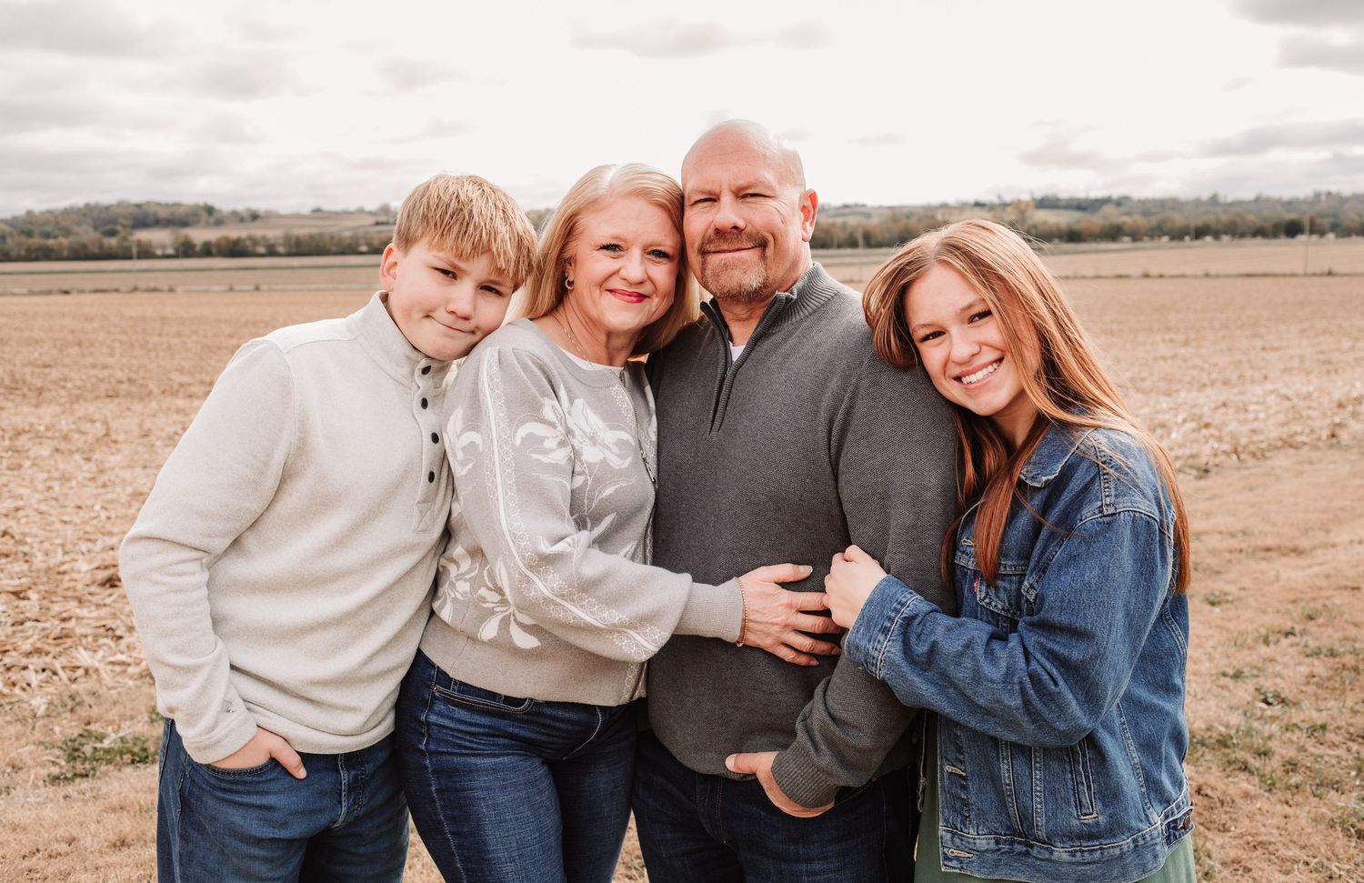 Smiling family of four huddled together in an open field, dressed in casual fall attire.
