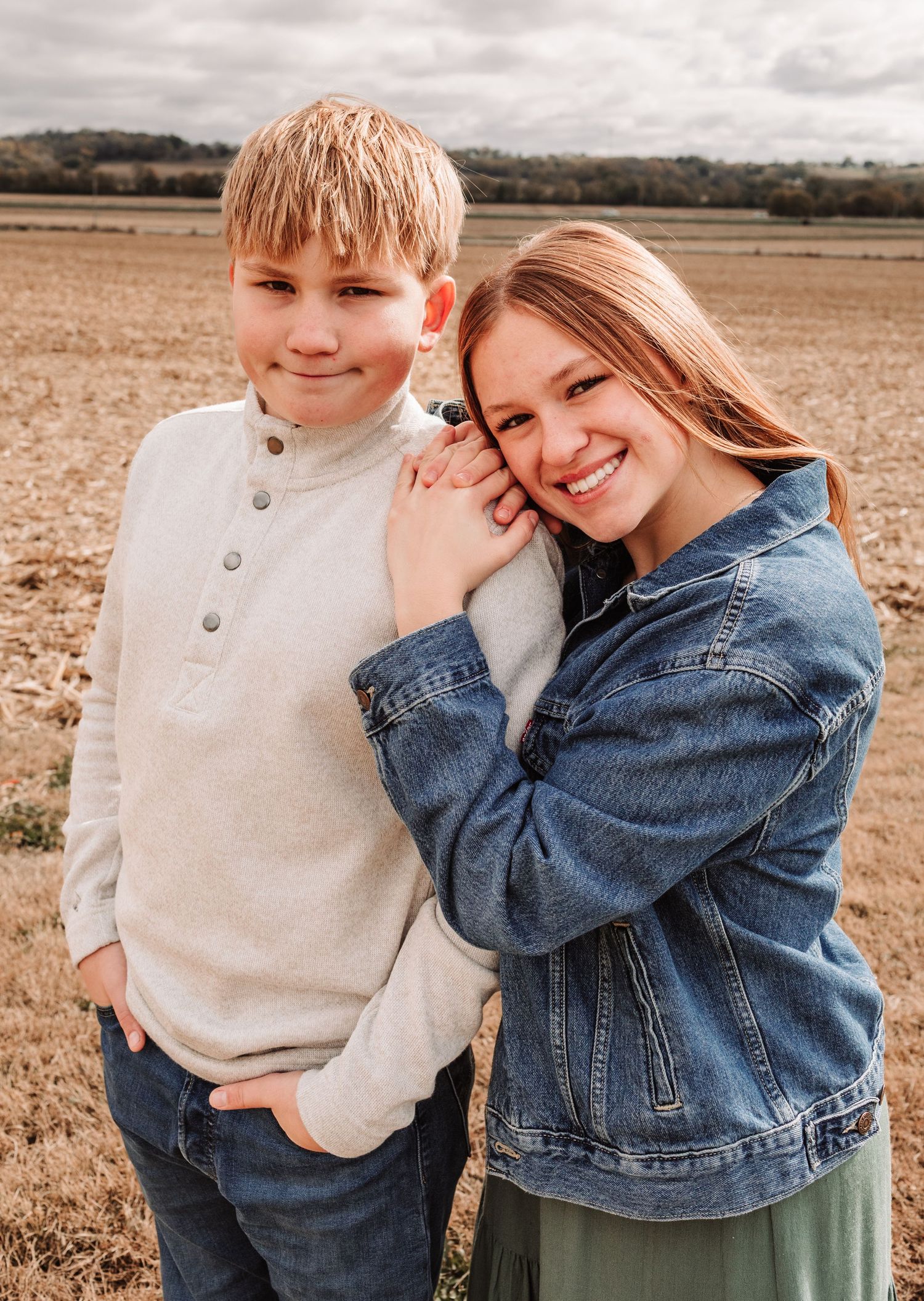 Teen girl leans on younger brother as they smile together in an open autumn countryside field.