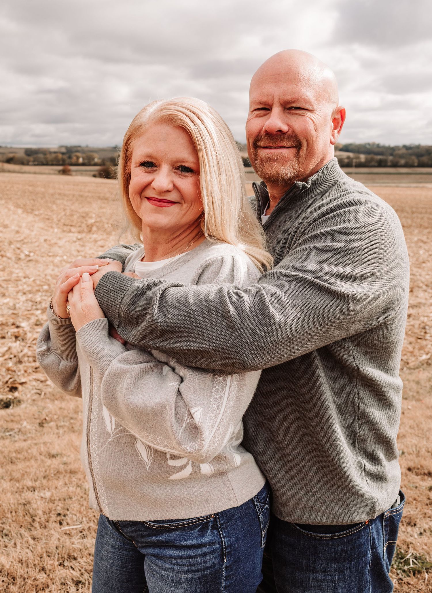 Smiling couple embracing in a harvested field, woman in light sweater and man in gray pullover.