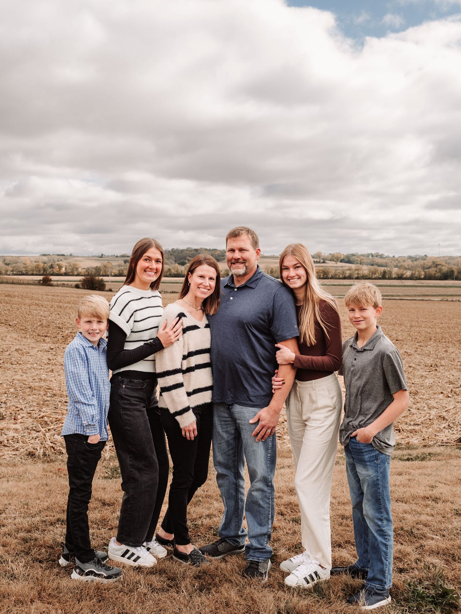 Family of six smiling together in a fall harvested field under a cloudy sky.