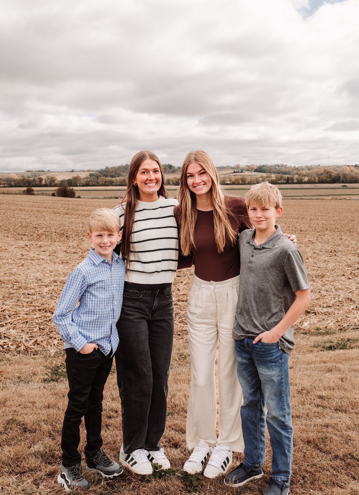 Four siblings posing together in a harvested field, two girls and two boys smiling outdoors.