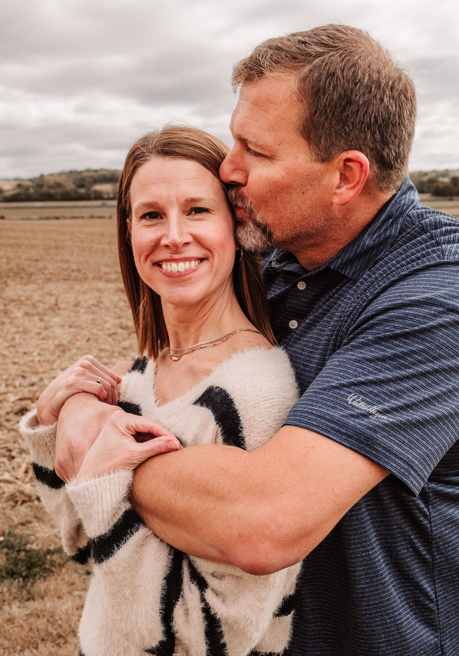 Couple embracing in a farm field, man kissing woman's cheek while she smiles warmly in a cozy striped sweater.