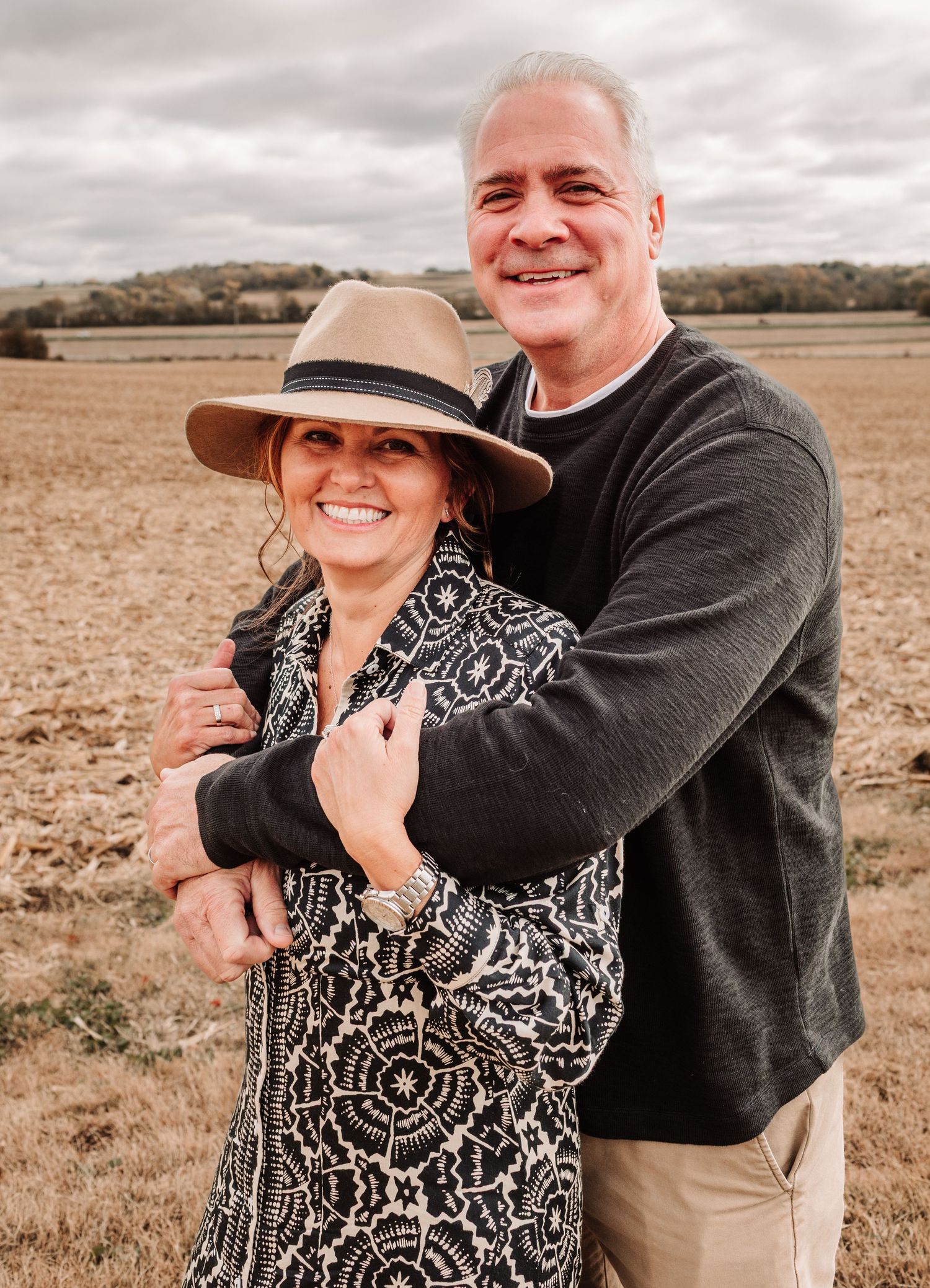 Smiling couple hugging outdoors in a harvested field, woman wearing a stylish wide-brim hat and patterned jacket.