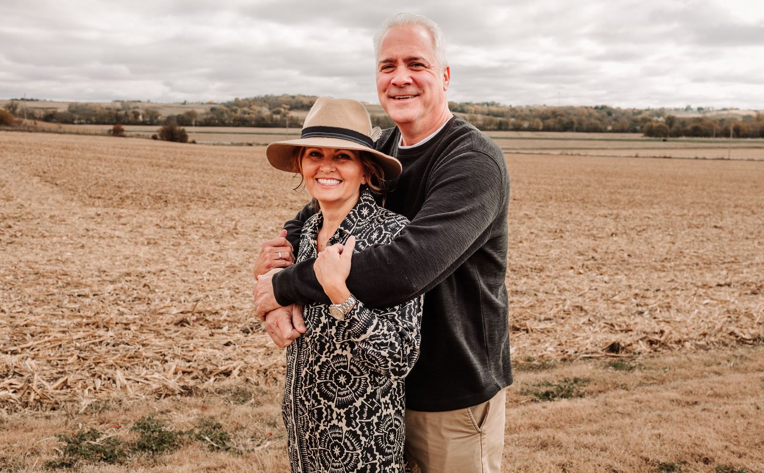 Happy couple embracing in an autumn farm field, woman in a wide-brim hat leaning into man in a dark sweater.