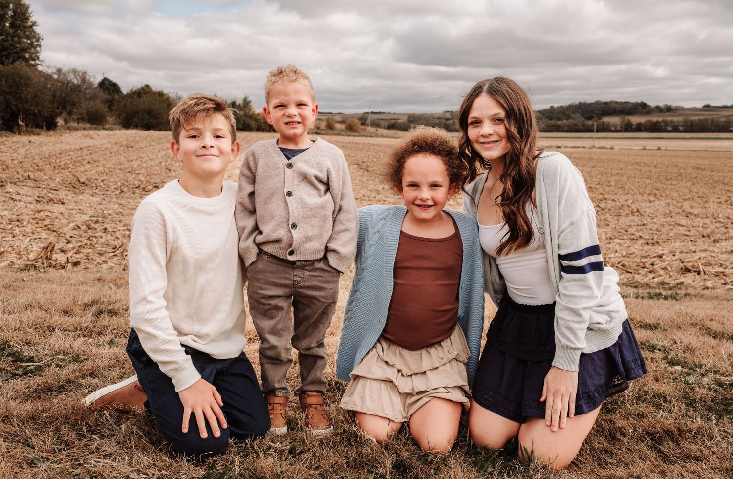 Four children smiling together in a golden farm field, posing casually in coordinated fall outfits.