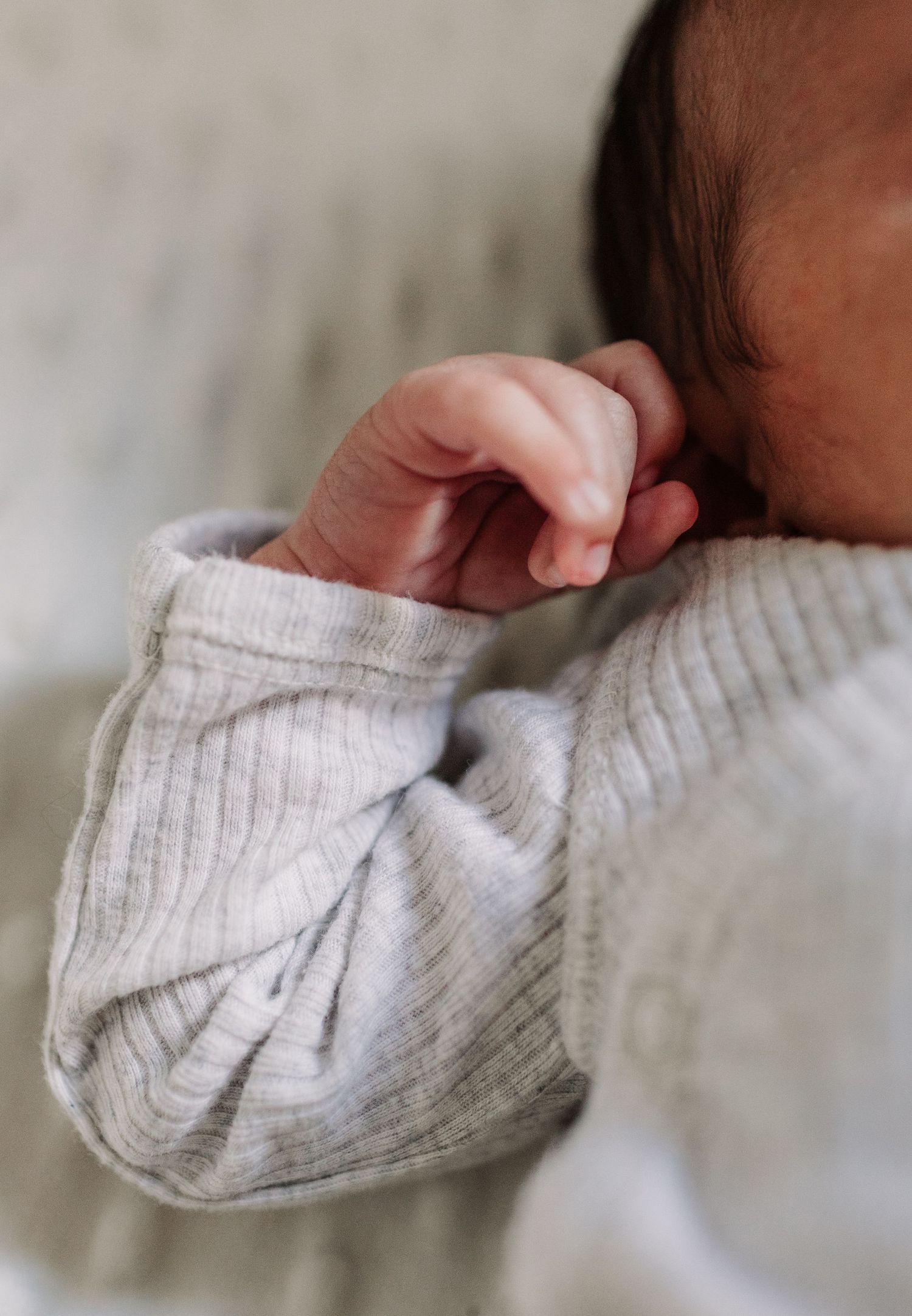Close-up of a sleeping newborn baby in a soft gray ribbed onesie with tiny fist raised near face.