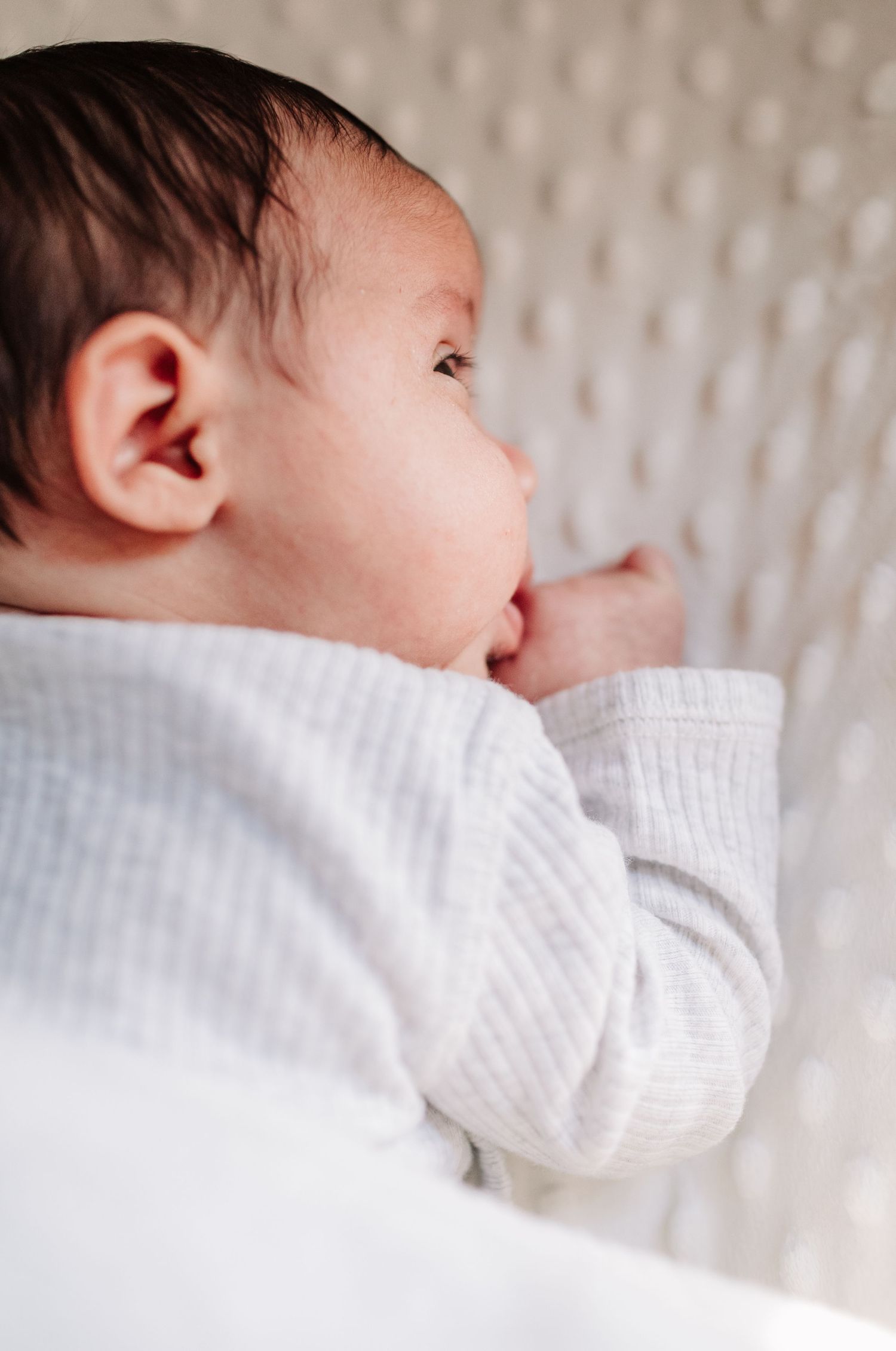 Newborn baby in a white ribbed onesie lying on side, gazing softly with hand near mouth.