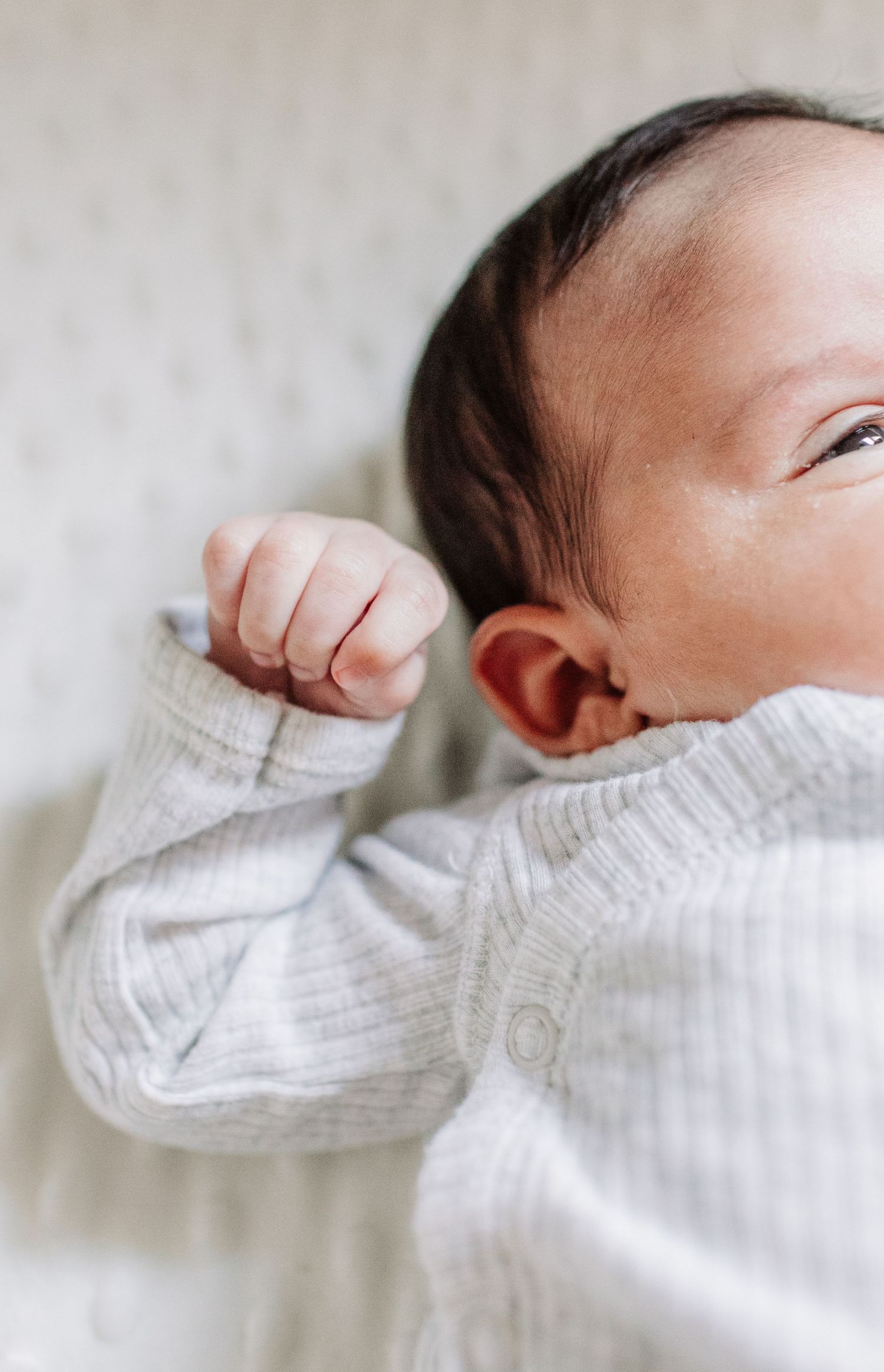 Peaceful newborn baby in a gray onesie lying down with tiny fist raised near face.