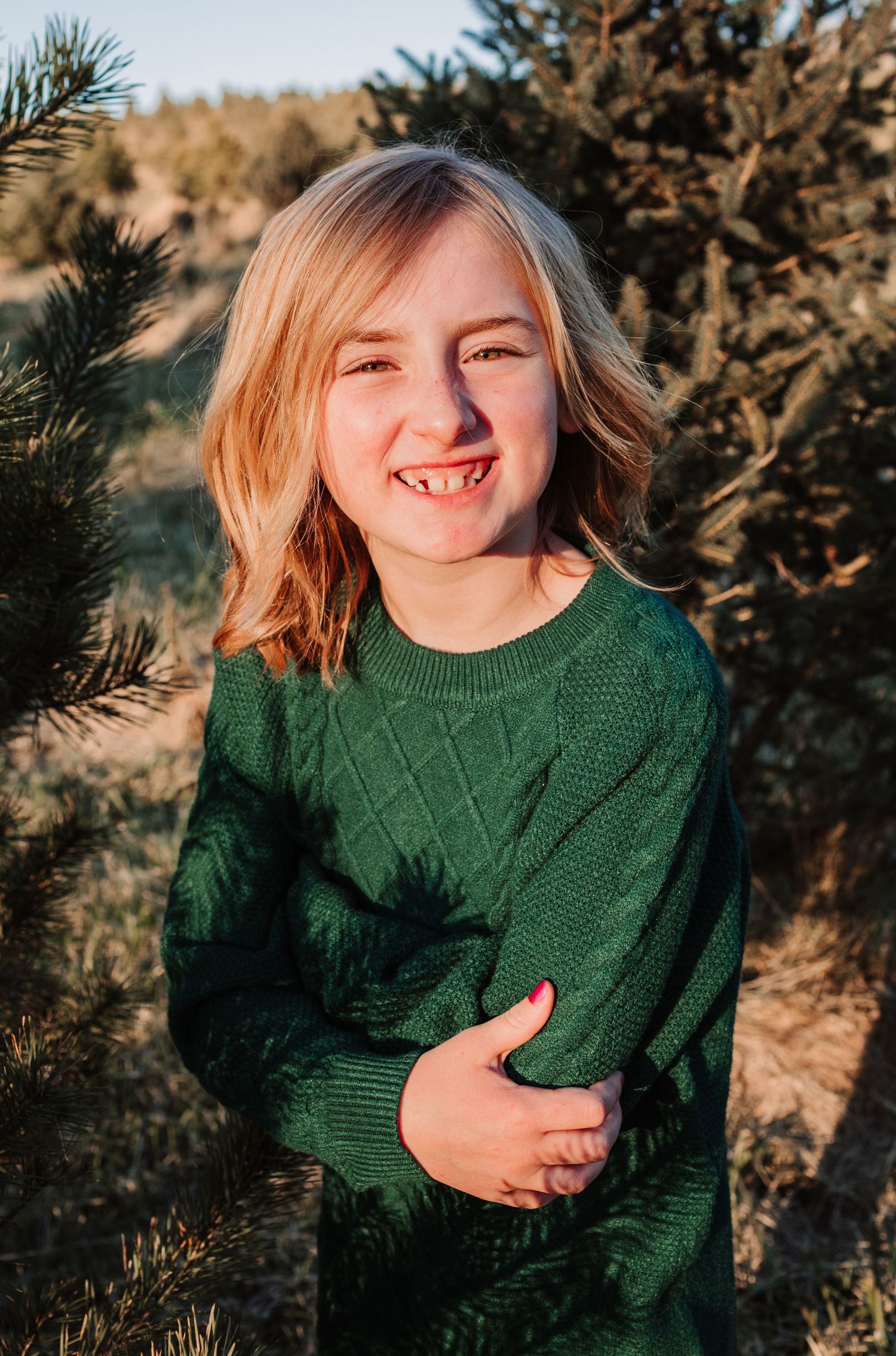 Cheerful blonde child in green sweater smiling and hugging herself at a Christmas tree farm.