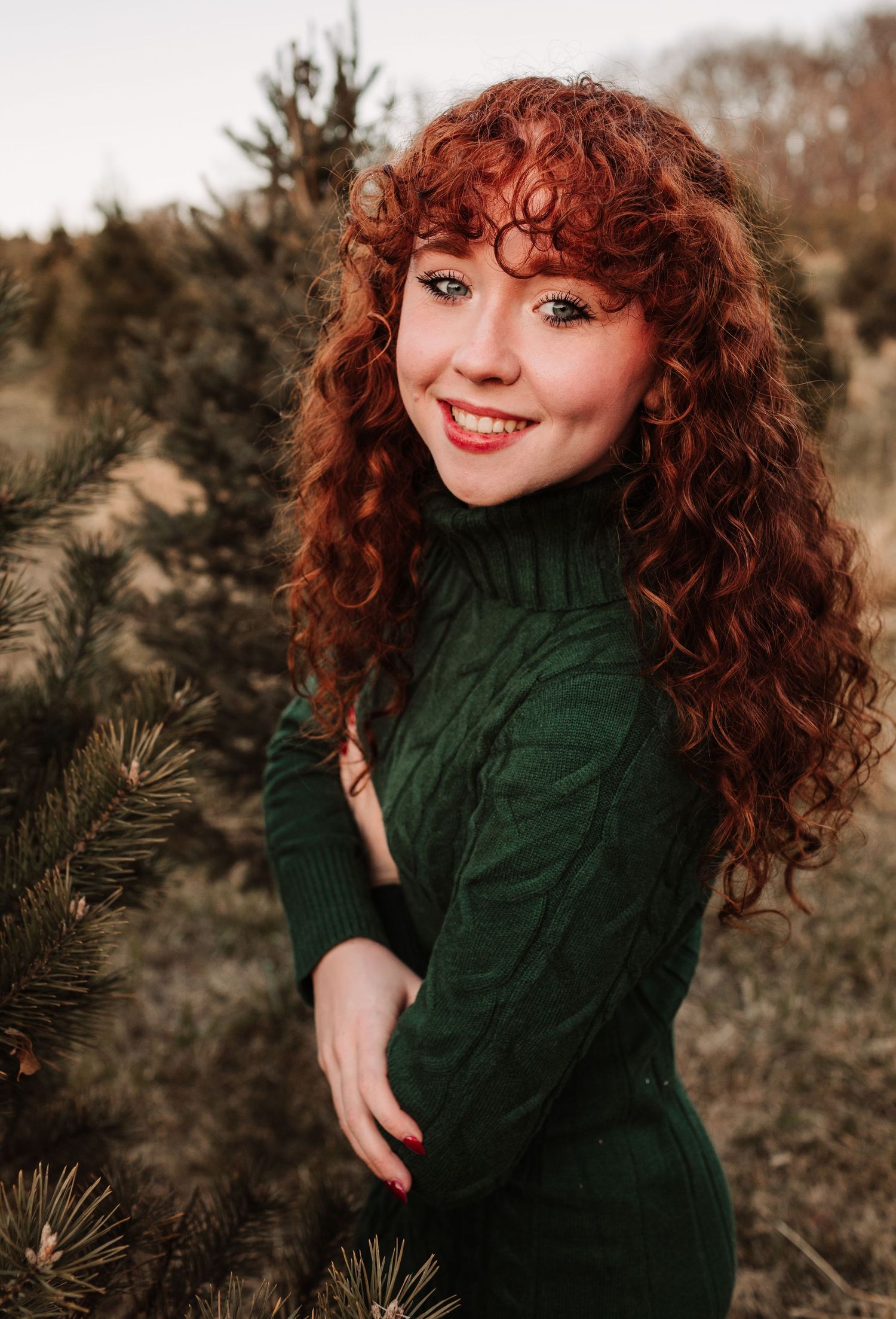 Beautiful young woman with curly red hair smiling in a green sweater at a Christmas tree farm.