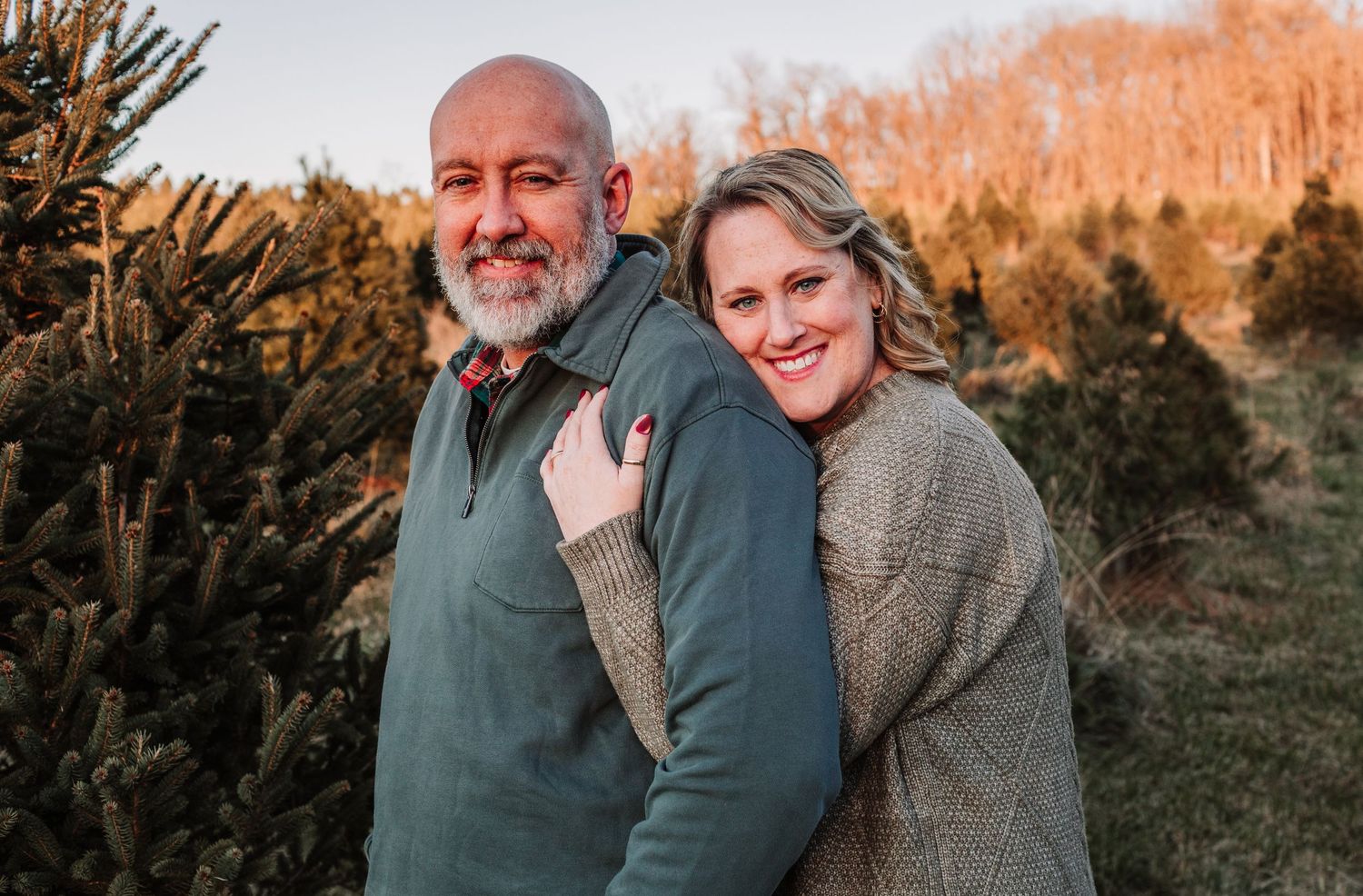 Smiling couple posing together in front of lush green Christmas trees at a tree farm.
