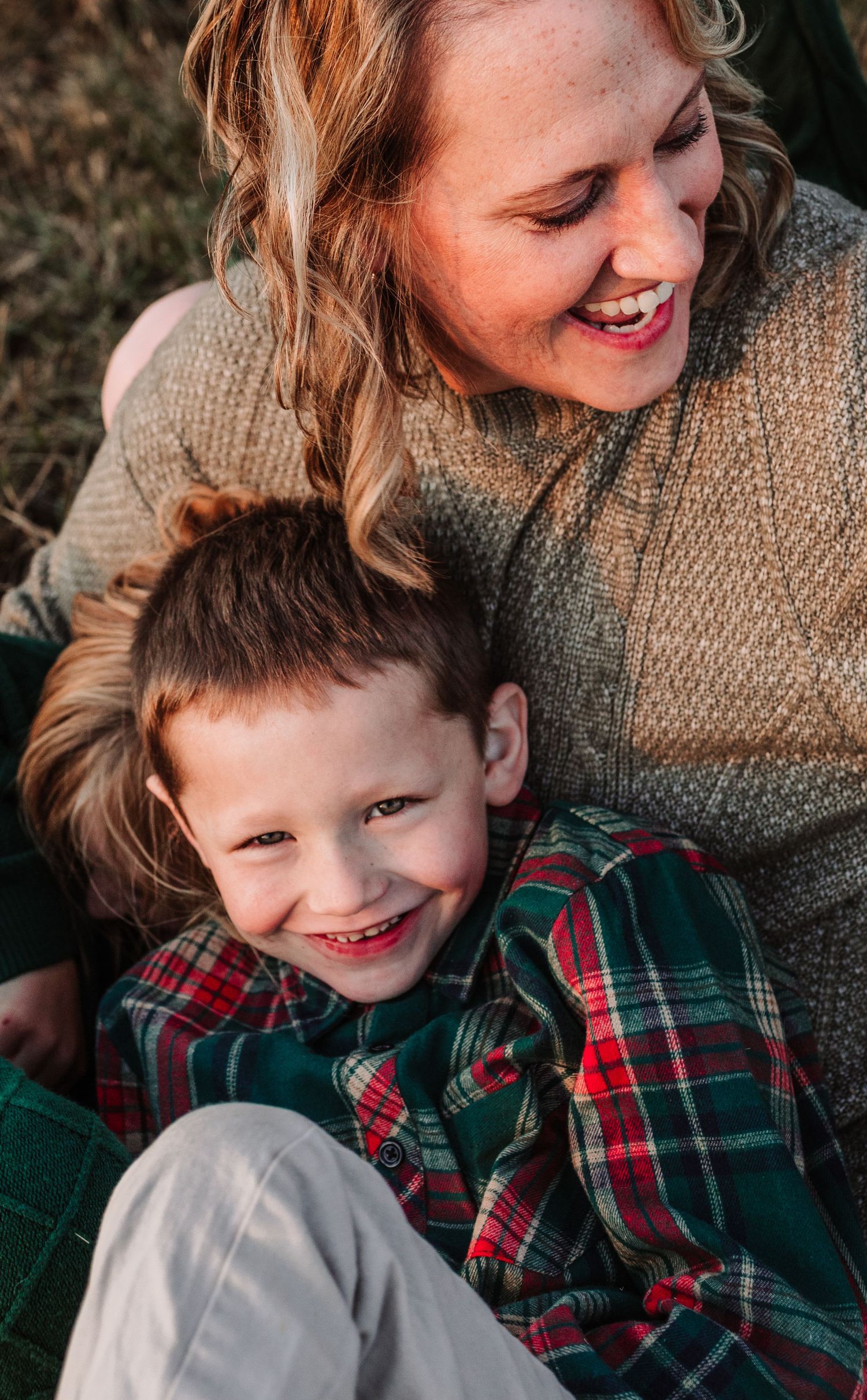 Smiling mother and young boy in red plaid shirt share a joyful moment during an outdoor holiday family portrait.