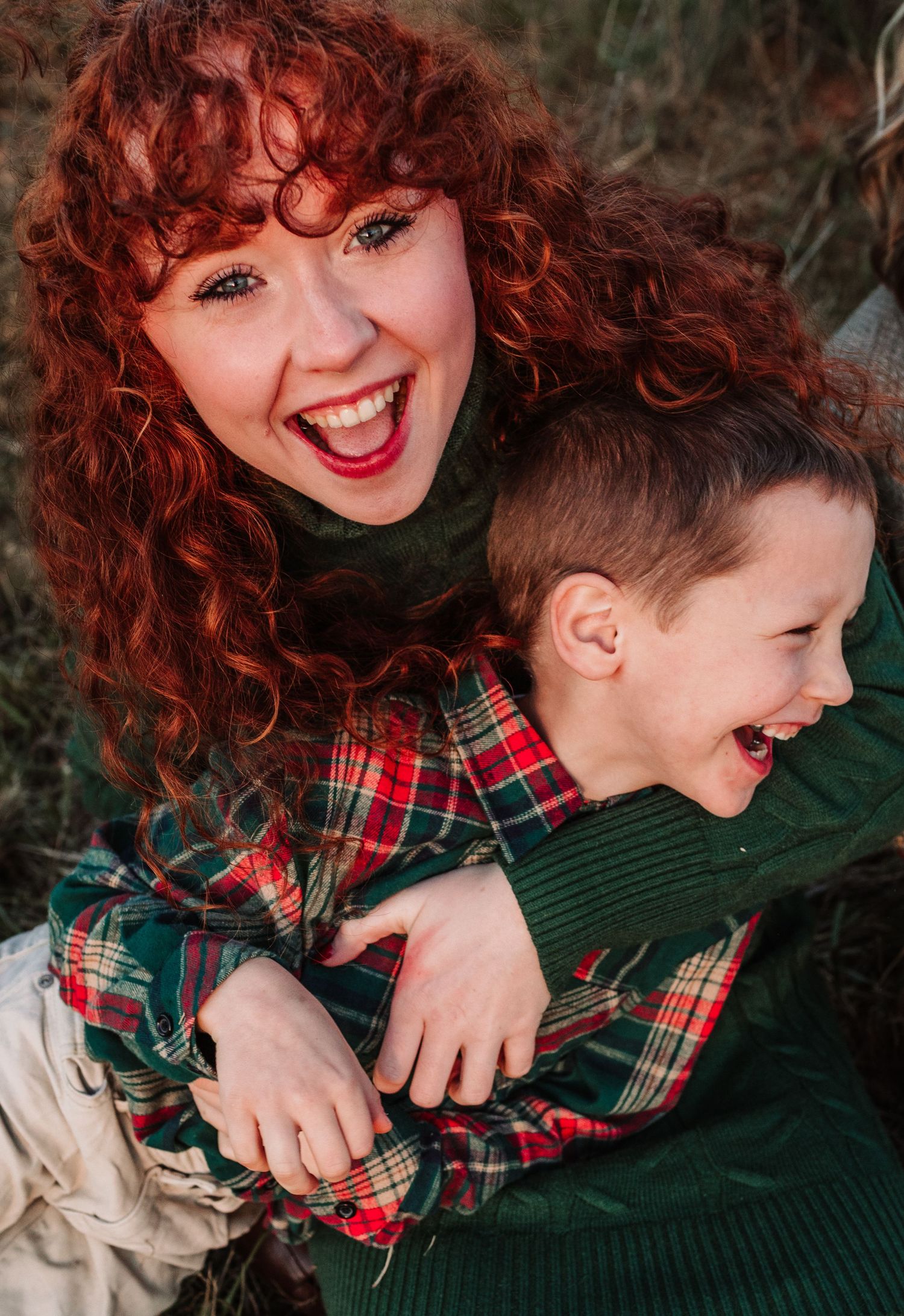 Laughing woman with curly red hair hugs a giggling child in plaid shirt during a festive outdoor photo shoot.
