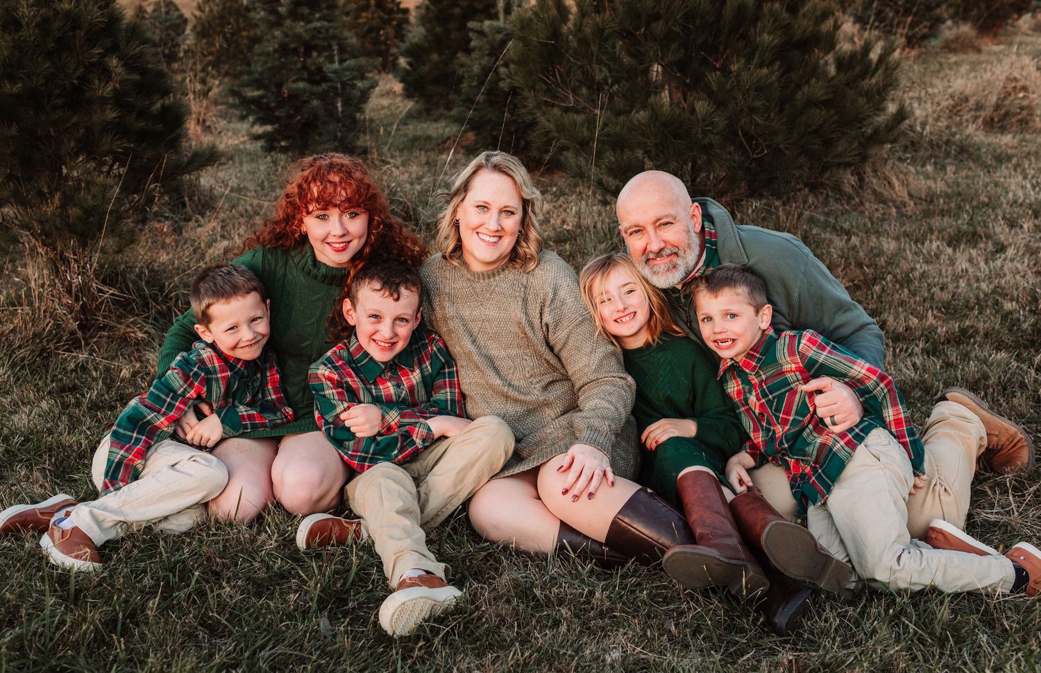 Happy family of seven poses together on grass in coordinated green and plaid holiday outfits near Christmas trees.