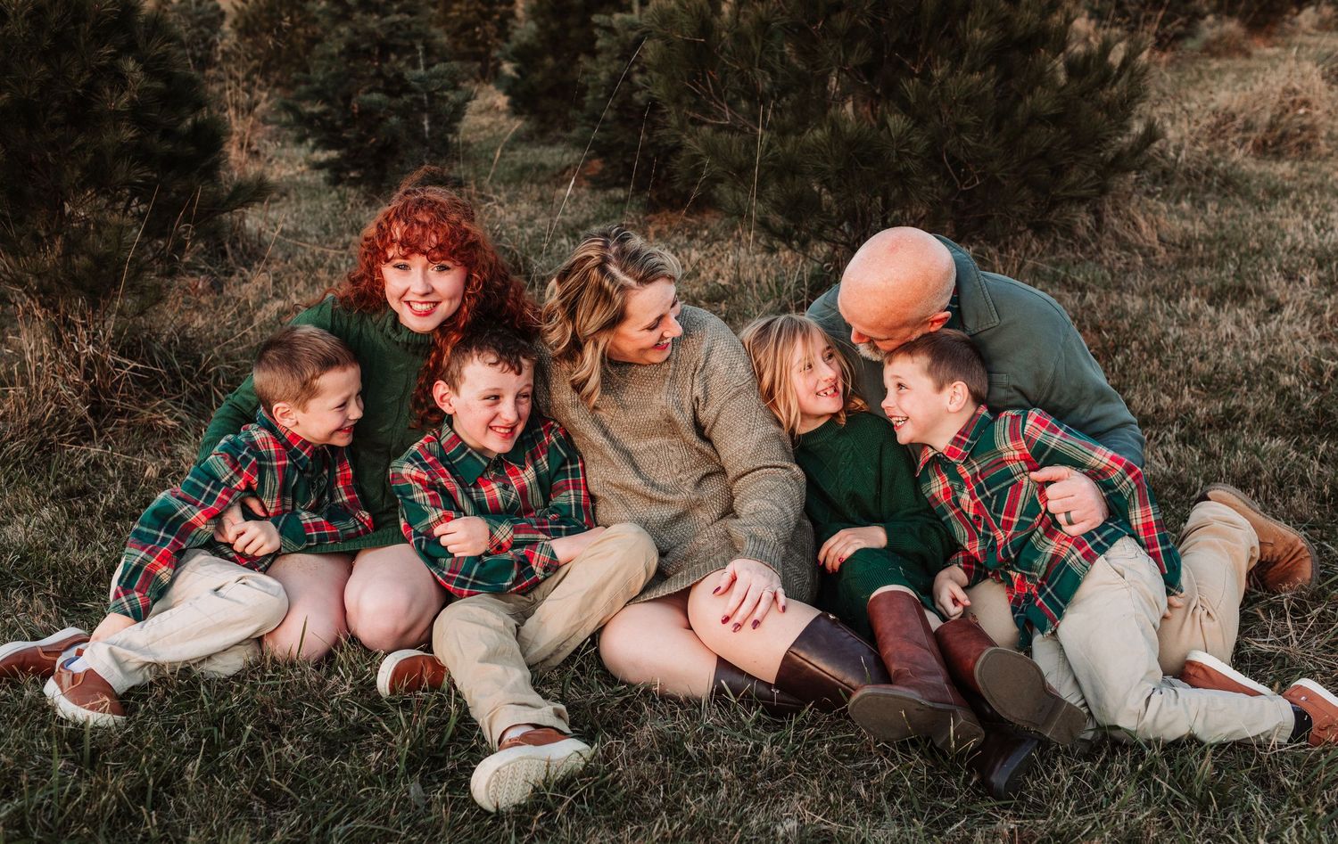 Joyful family of seven laughs and cuddles together outdoors in festive green and plaid holiday clothing.