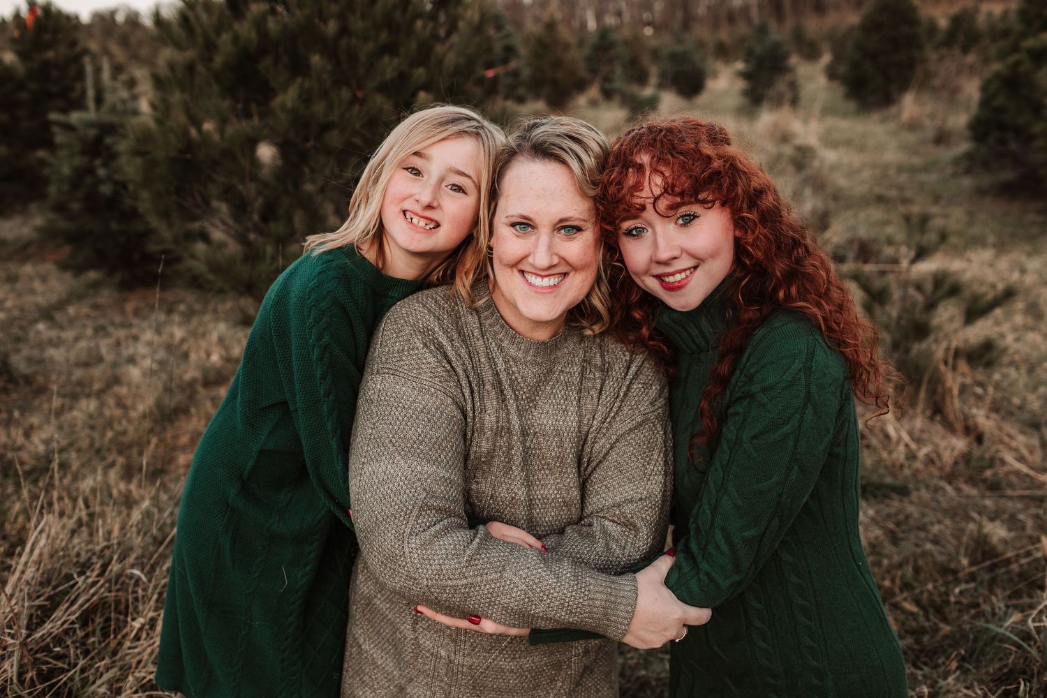 Three smiling women in green and neutral sweaters pose together outdoors during a festive family photo session.