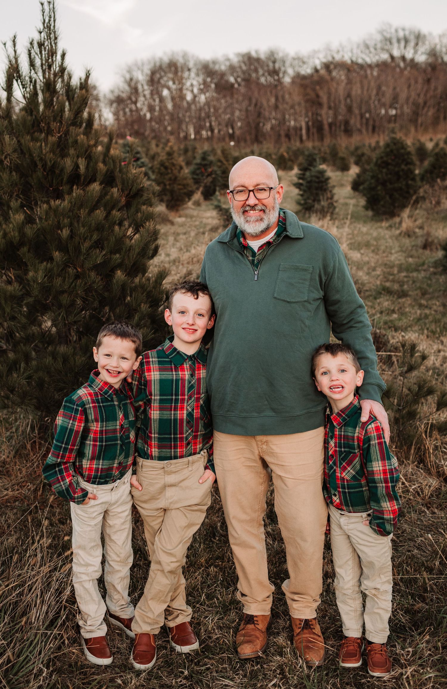 Father stands proudly with three young boys in matching red plaid shirts at a Christmas tree farm.