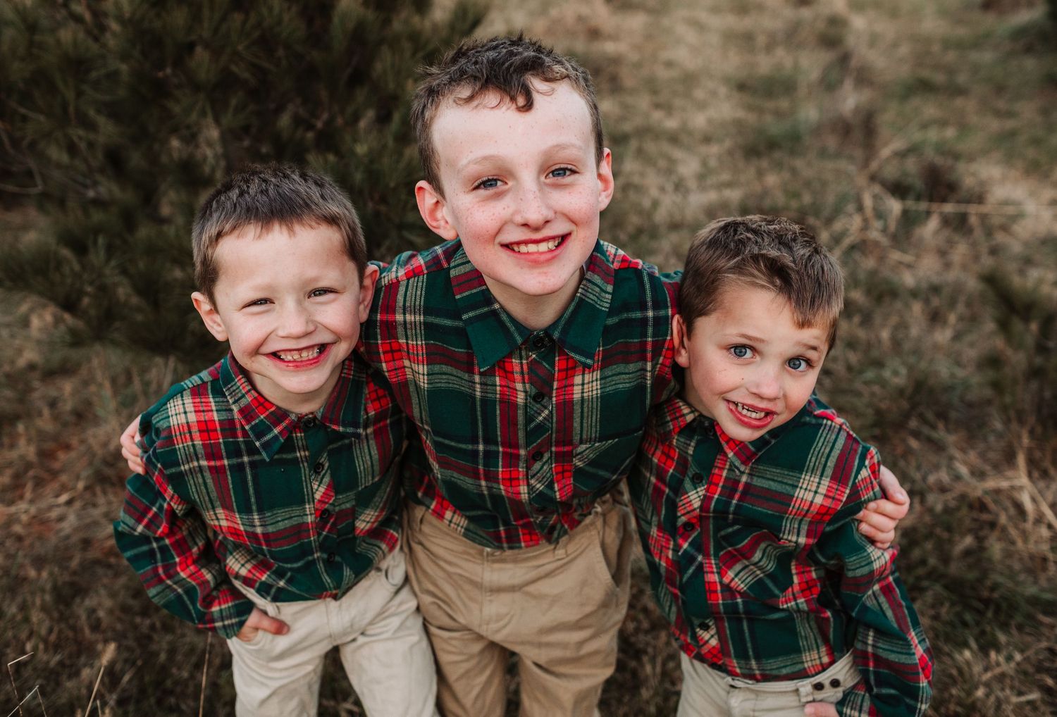 Three young brothers in matching red and green plaid shirts smile up at the camera during a holiday photo session.