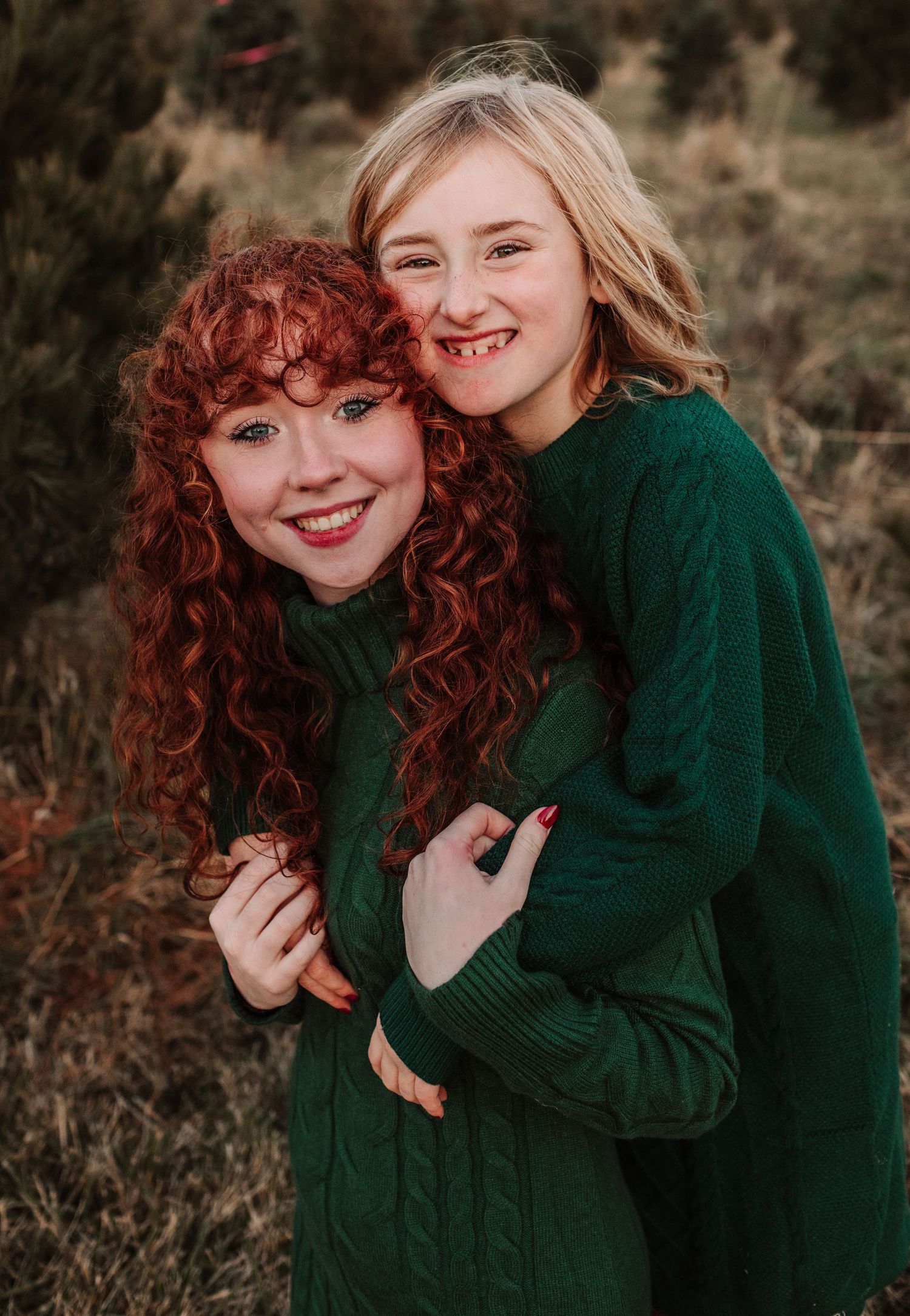 Two smiling girls in matching green cable-knit sweaters pose together outdoors at a Christmas tree farm.