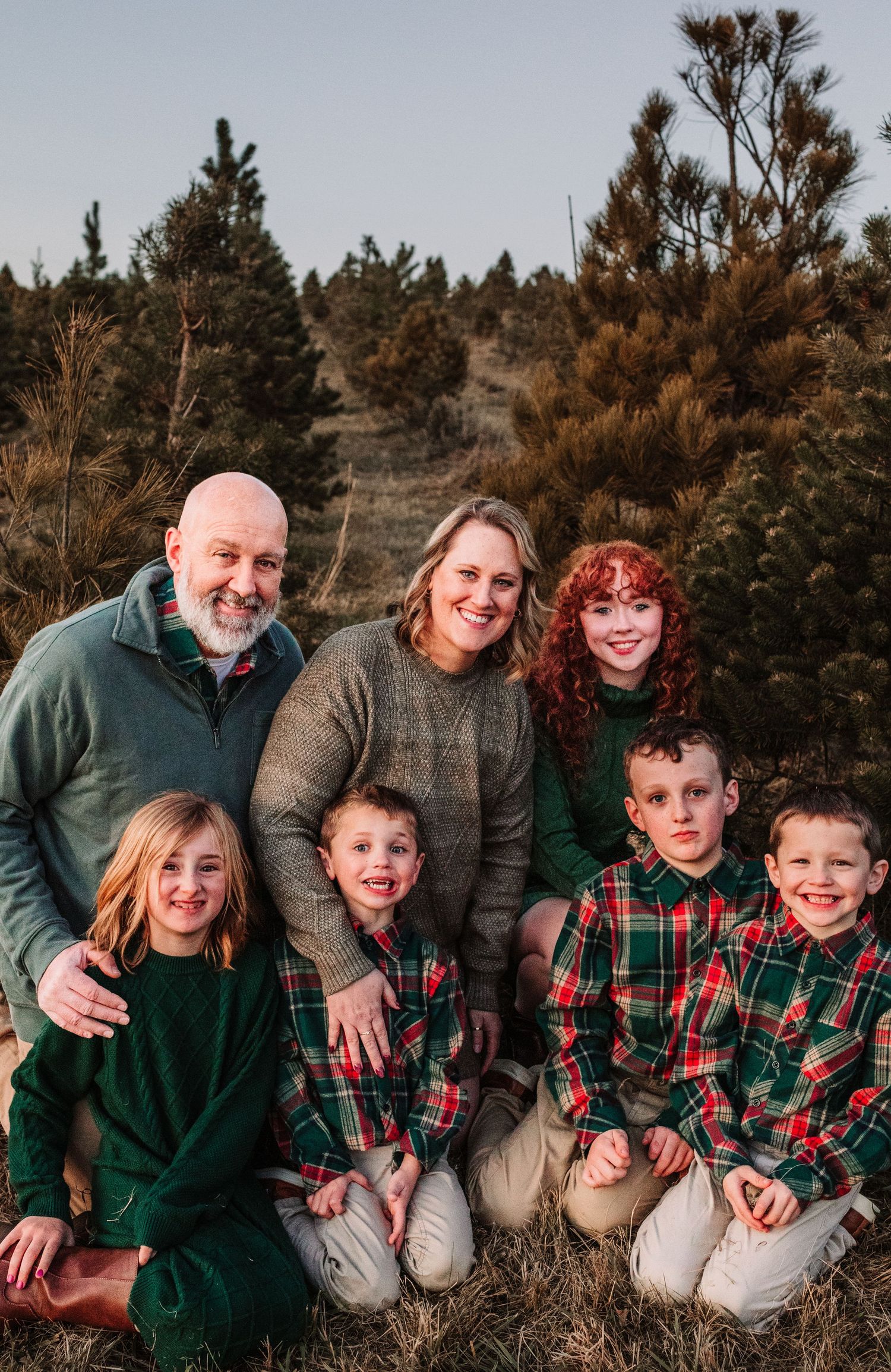 Family of seven poses together at a Christmas tree farm, wearing coordinating green and plaid holiday outfits.