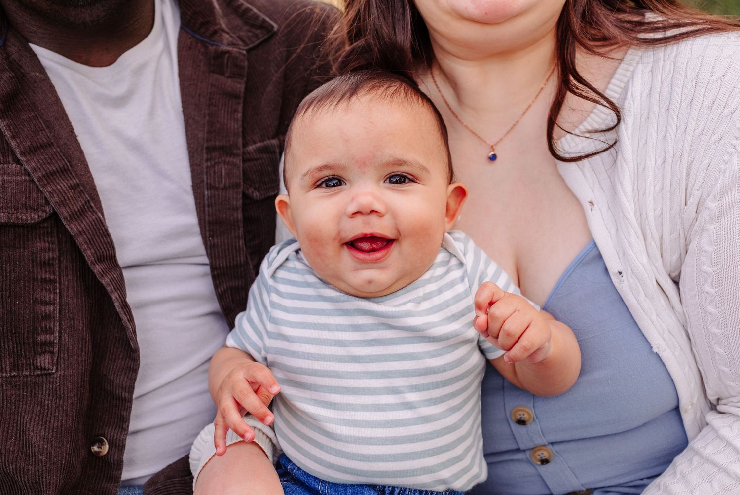 Smiling baby in striped onesie held by interracial parents outdoors.