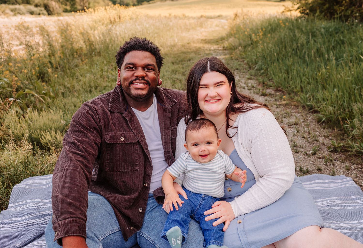 Happy interracial family with baby sitting on blanket in green field.
