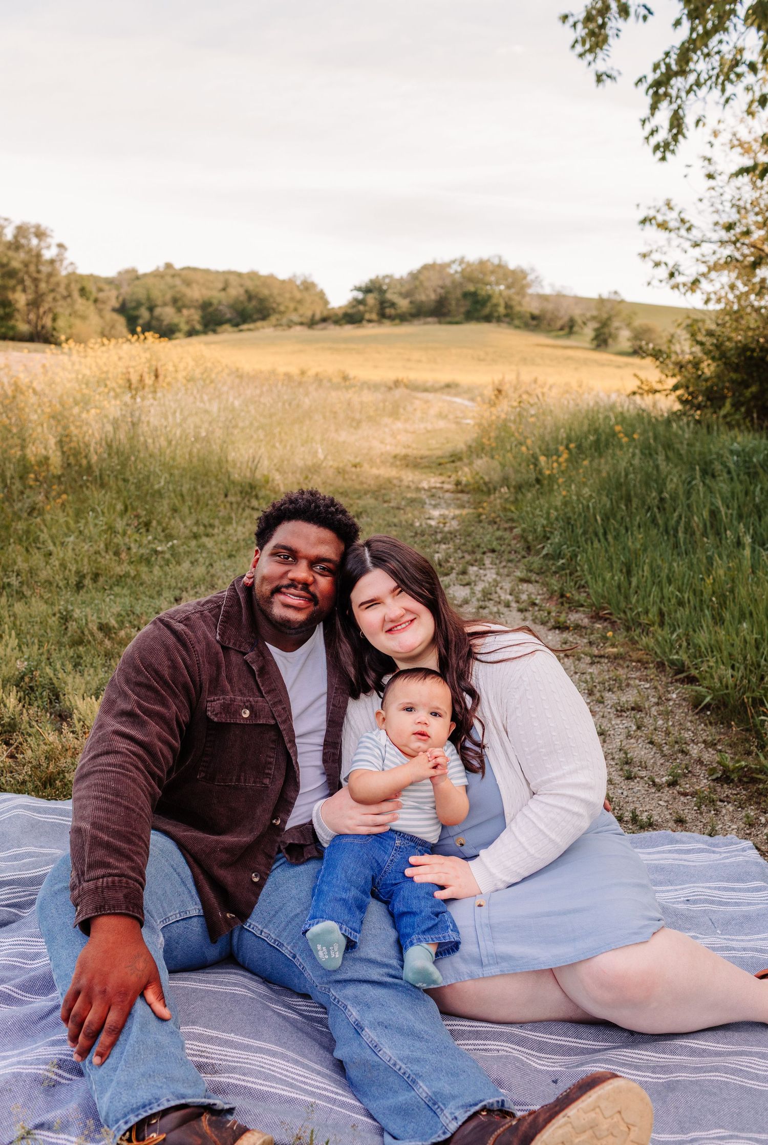 A happy family of three sits on a blanket in a scenic field, with parents smiling and holding their baby.