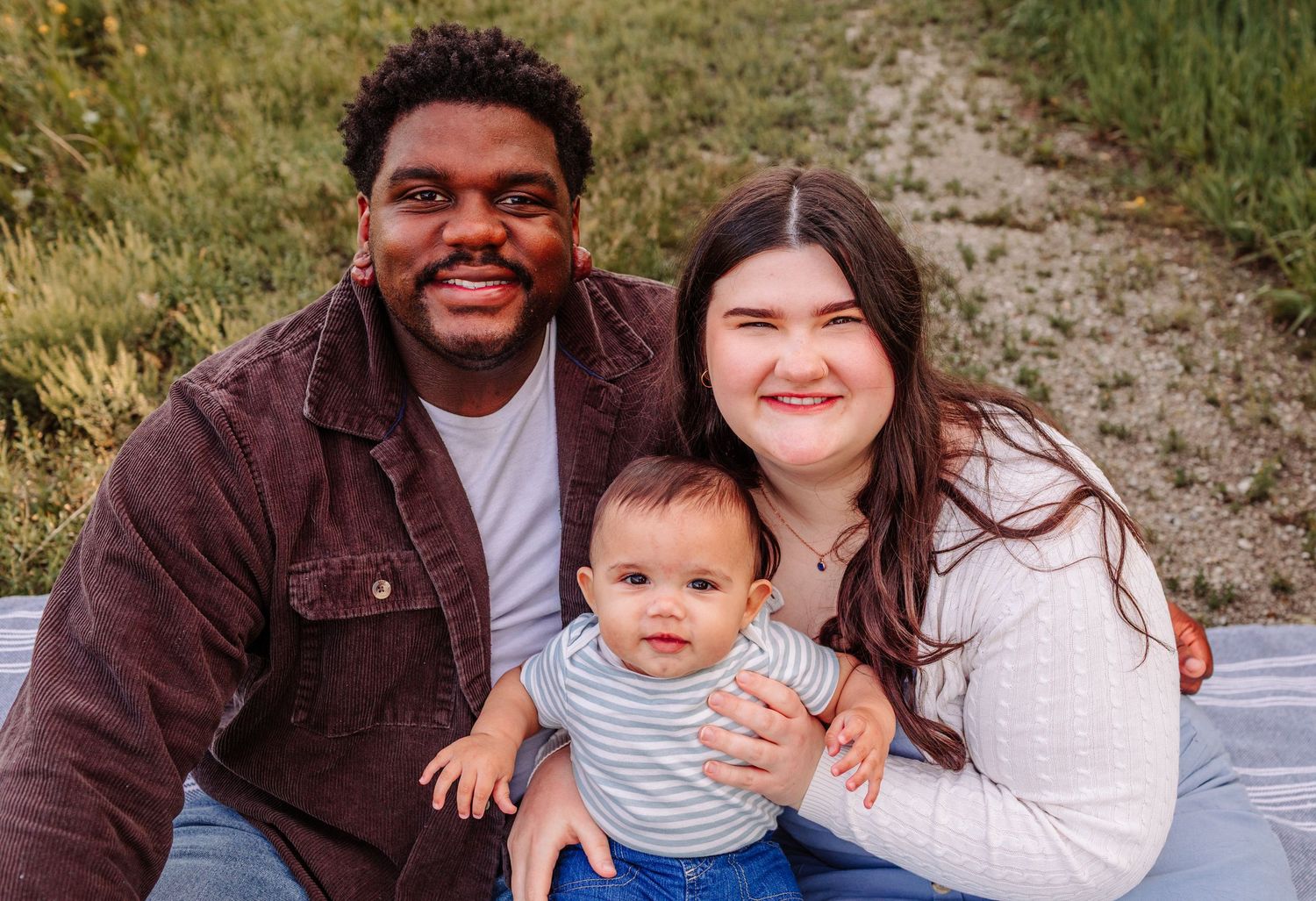 Happy interracial family with smiling parents and adorable baby sitting outdoors in a green field during golden hour.