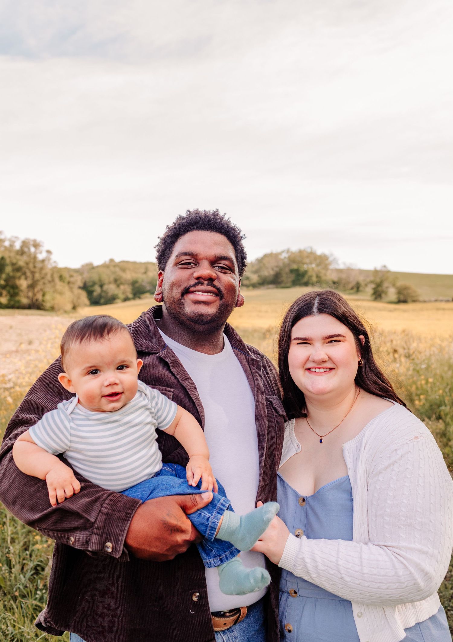 Smiling interracial couple holding baby in sunny outdoor field.