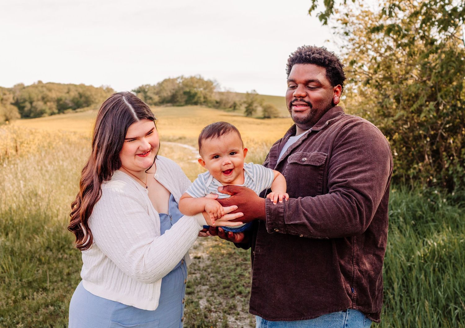 Happy interracial family with smiling baby outdoors in golden field during sunset family photo session.