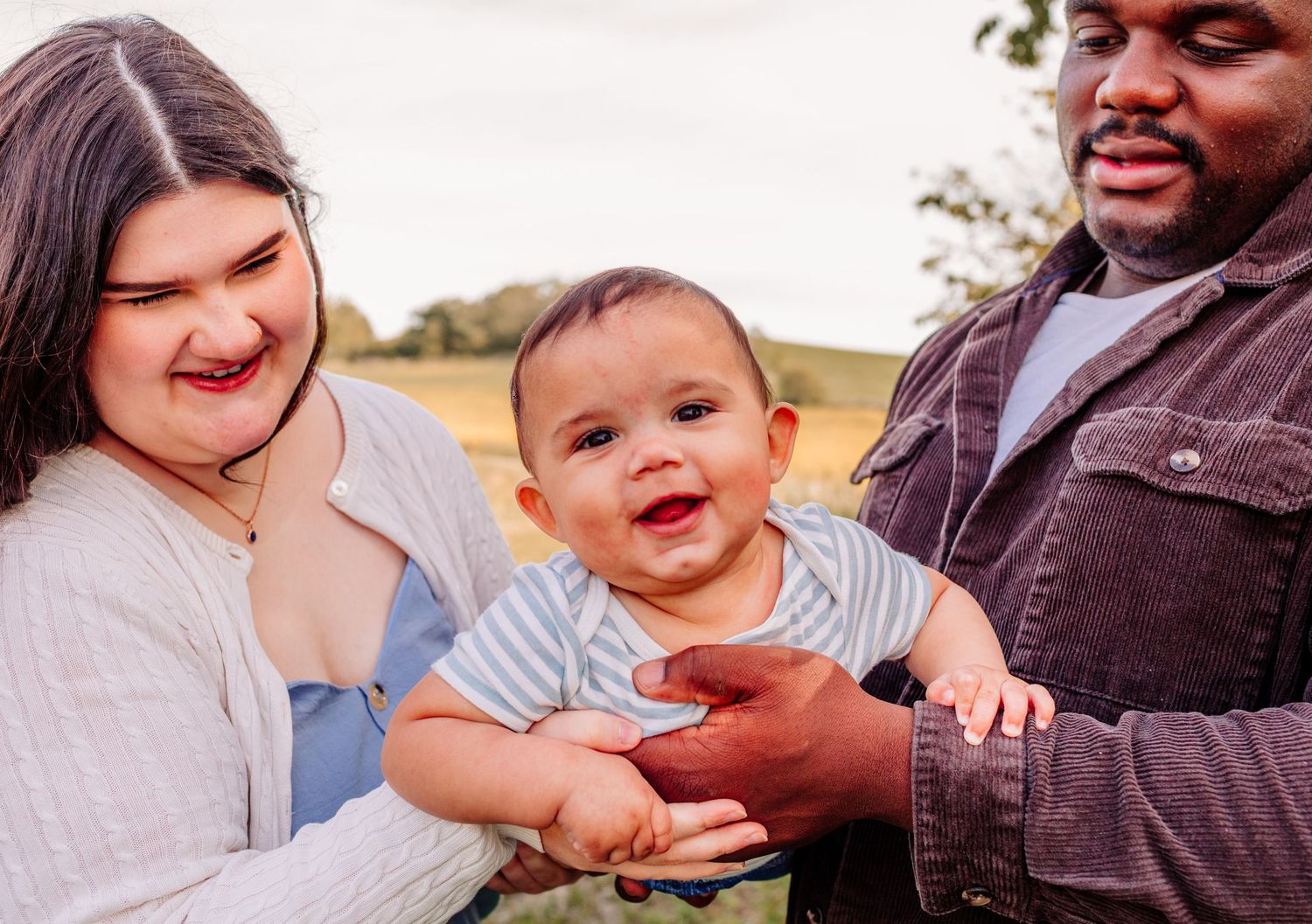 Happy mixed-race family with smiling baby outdoors in a green field, parents holding infant together.